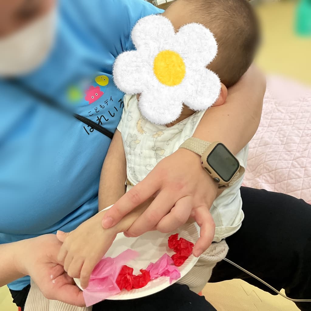 はれいろの支援員に抱っこされた乳児が、手を伸ばして赤やピンクの花紙に触れながら工作活動に参加している様子