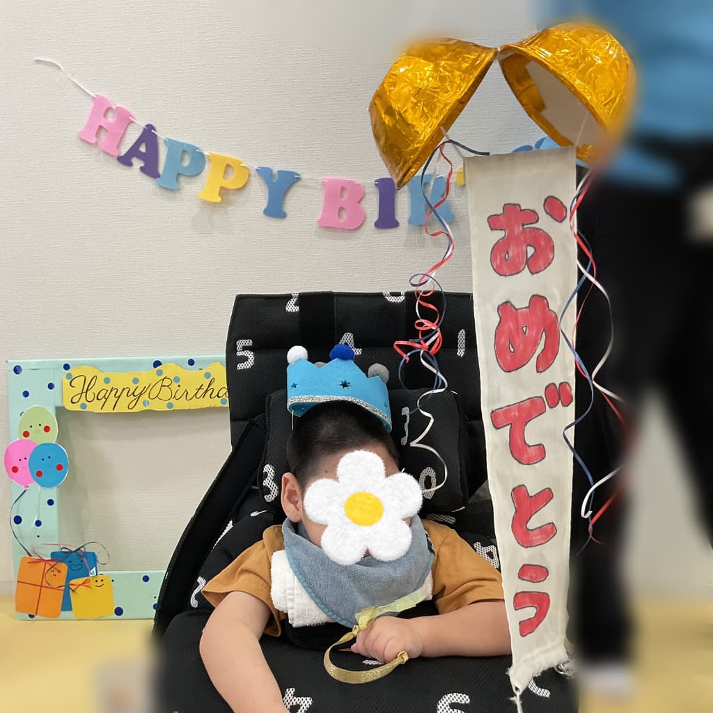 青い王冠をかぶった子どもが、金色のくす玉や「おめでとう」と書かれた飾りの前で誕生日のお祝いを受けている様子