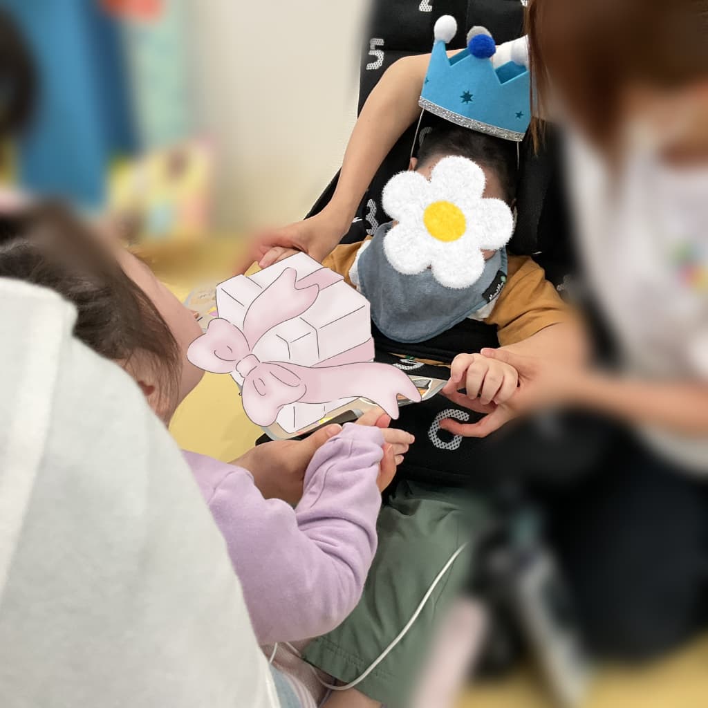 王冠をかぶった子どもが、周囲の子どもたちや支援者に囲まれながら、リボン付きのプレゼント型カードを受け取って誕生日をお祝いされている様子