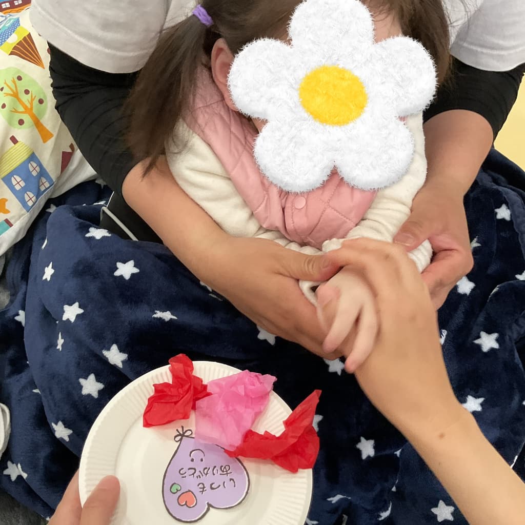支援員が子どもの手を支えながら、花紙を丸めて貼り、「いつもありがとう」と書かれたハート型の母の日工作を一緒に作っている様子