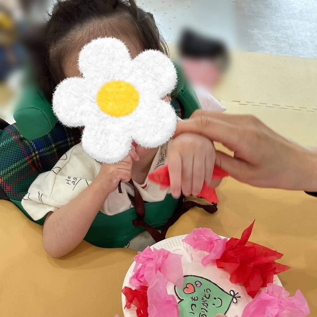 支援員に手を添えてもらいながら、子どもが赤い花紙をちぎって貼ろうとしている。「いつもありがとう」と書かれたハート型の母の日工作を作っている様子