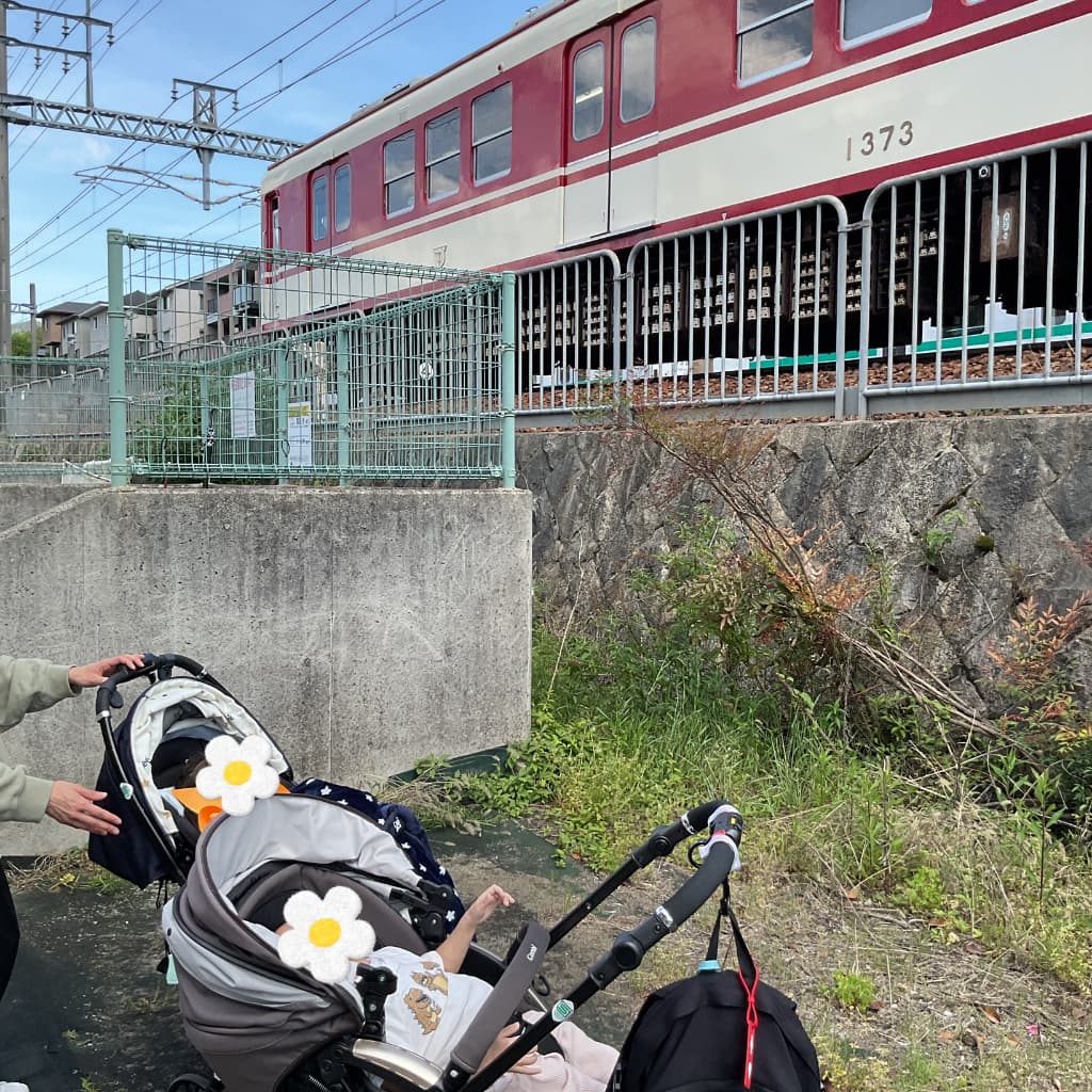 ベビーカーに乗った子どもたちが線路沿いで赤い電車を見上げなている様子