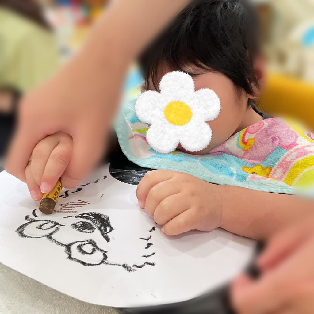 子どもが支援者に手を添えてもらいながら、白い紙に描かれた動物のイラストへ黒いクレヨンで色を重ねている様子