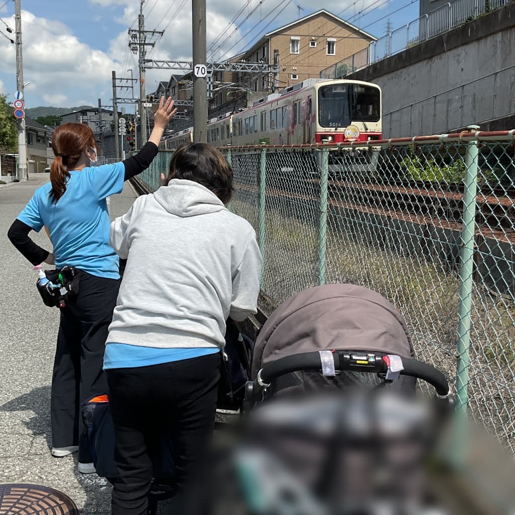 支援員2人とベビーカーに乗った子どもたちが線路沿いで通過する電車に向かって手を振っている様子
