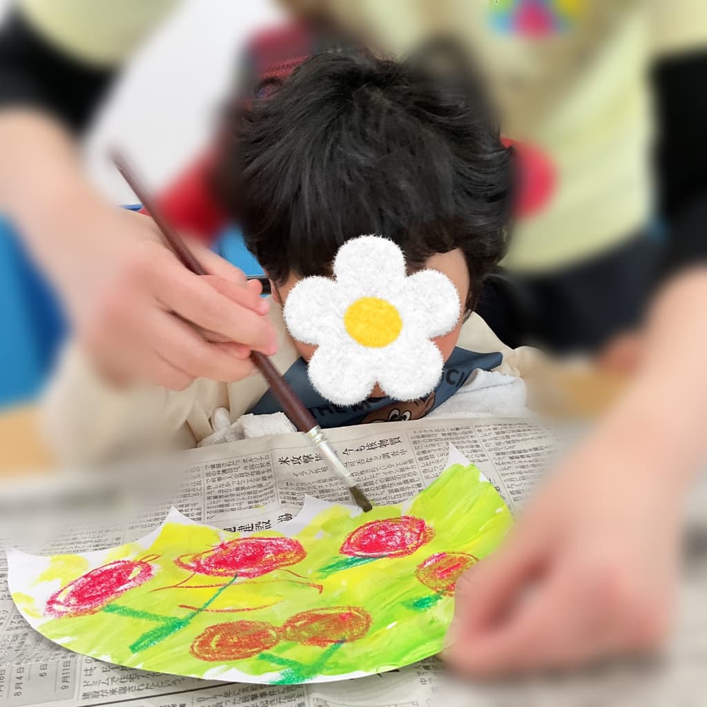 子どもが支援者と一緒に筆を持ち、赤い花の絵が描かれた作品に黄緑色の絵の具を塗りながら制作活動に取り組んでいる様子