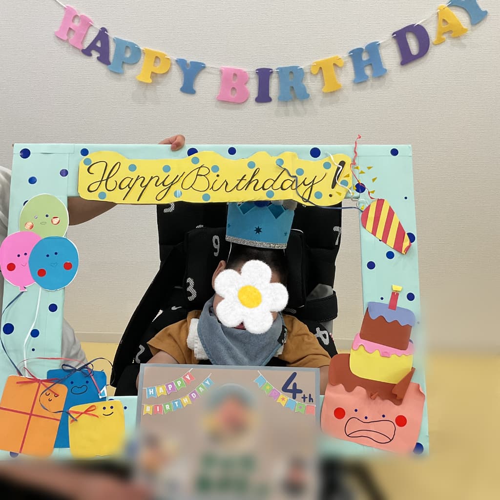 誕生日を迎えた子どもが、青い王冠をかぶり、「HAPPY BIRTHDAY」の飾りや手作りフレームに囲まれて記念撮影をしている様子