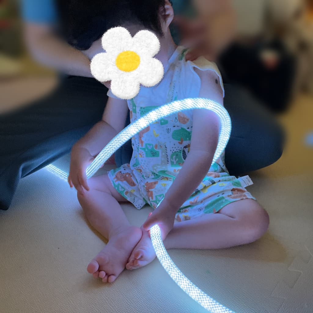 薄暗い室内で、支援者に身体を支えられながら座った子どもが、白く光るロープ状のライトをもって遊んでいる様子