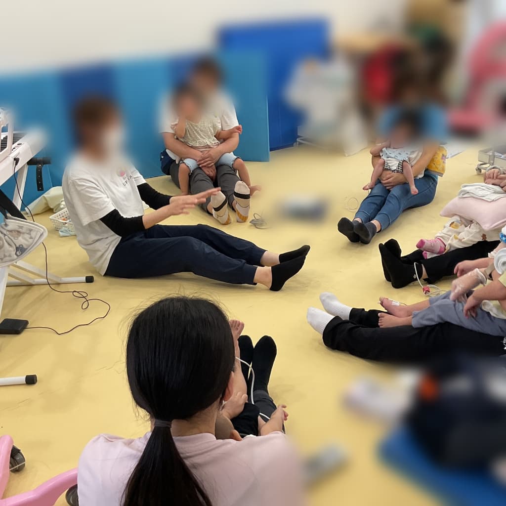 室内で、大人と子供がペアとなってが円になるように床に座り、中央に向かって集まって活動をしている様子。