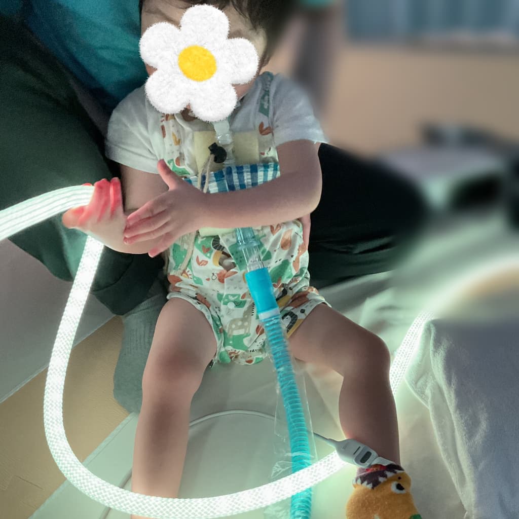 薄暗い室内で、支援者に支えられて座る子どもが、手のひらで白く光るロープ状のライトに触れている様子。胸元から呼吸器のチューブが伸び、足元まで続いている