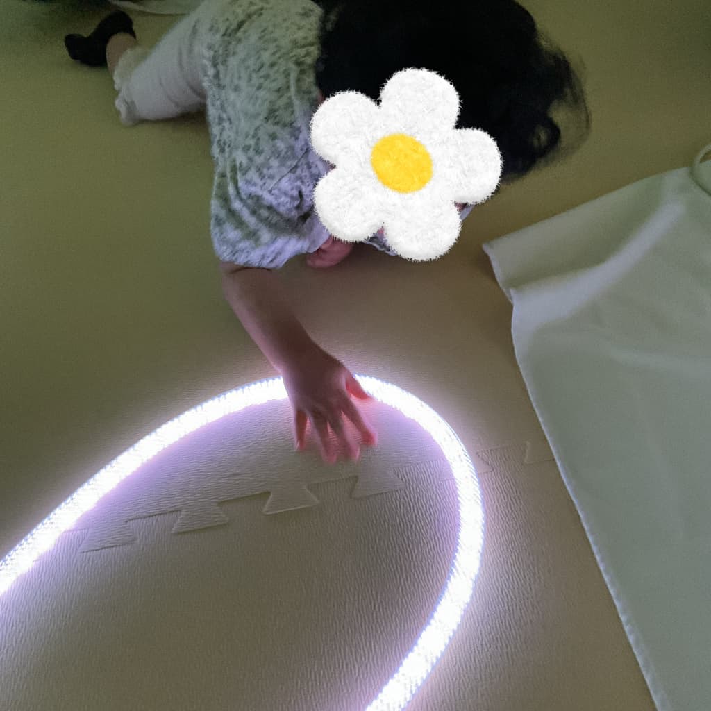 薄暗い室内で、床にうつ伏せに近い姿勢になった子どもが、床に大きく弧を描いて置かれた白く光るロープ状のライトへ片手を伸ばして触れている様子