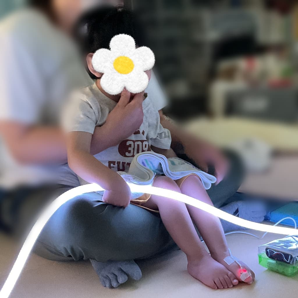 薄暗い室内で、支援者の膝に座った子どもが、膝の前を通る白く光るロープ状のライトに手を添えている様子