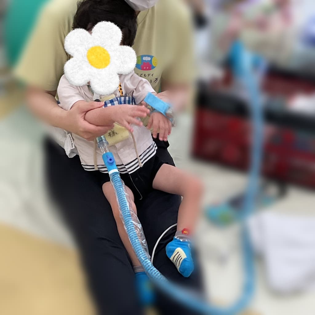 室内で、子どもが大人に抱っこされながら、敵日につけた鈴を鳴らしている様子。呼吸器用の青いチューブが、首元から足元まで続いている。