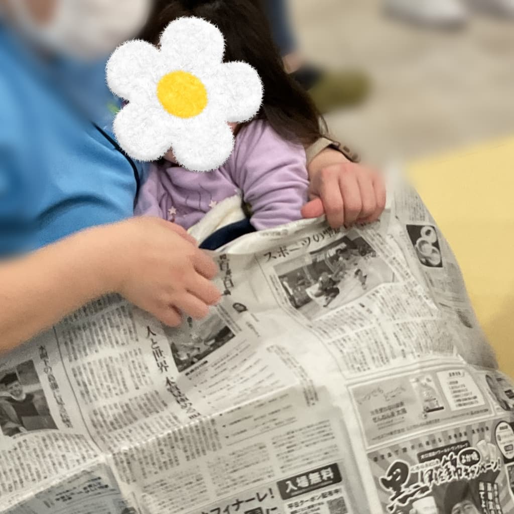 室内で、大人に抱きかかえられた子どもが新聞紙を両手で持ち、膝の上に広げている様子