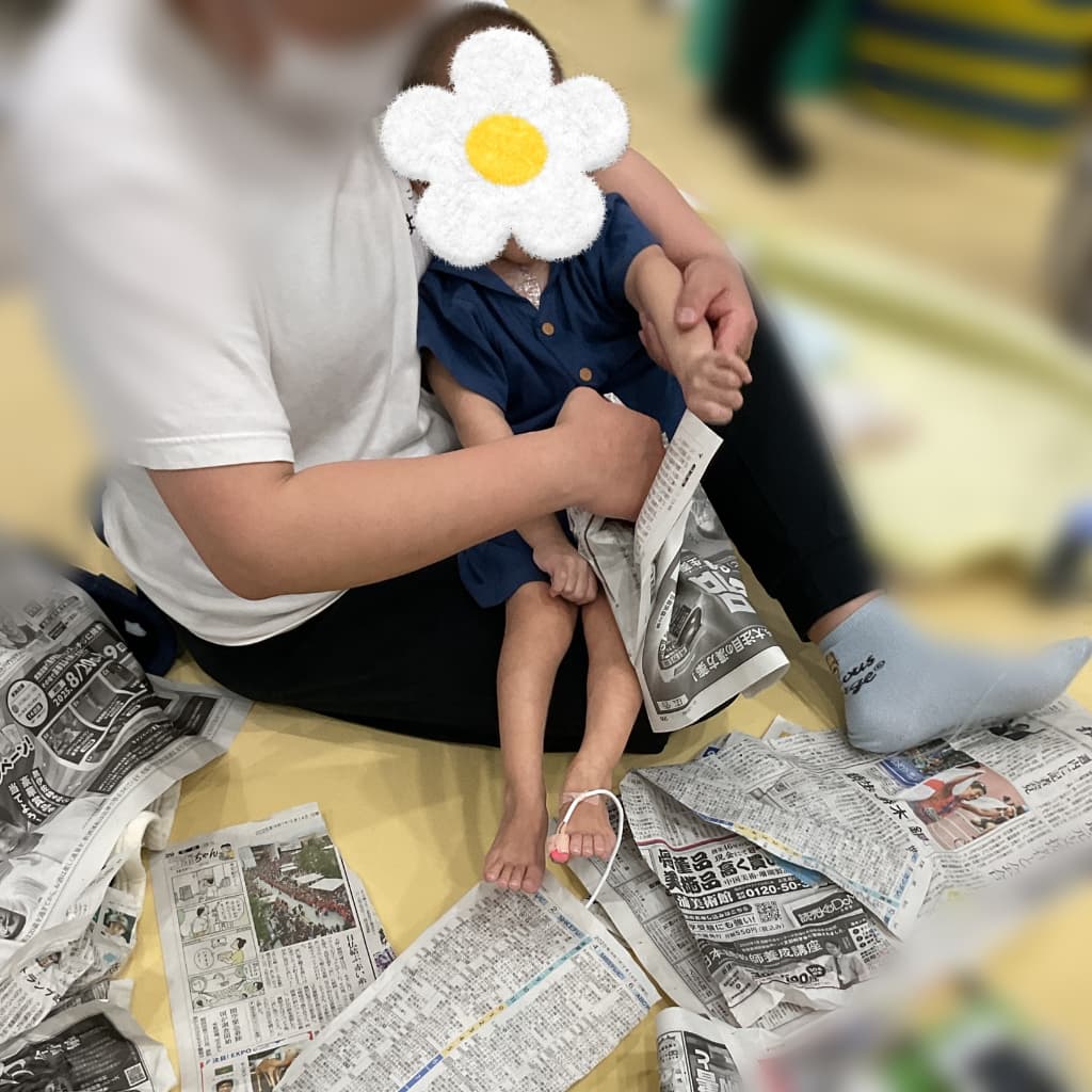 室内で、大人に抱っこされた子どもが、床に広げられた新聞紙の上で手を伸ばしている様子