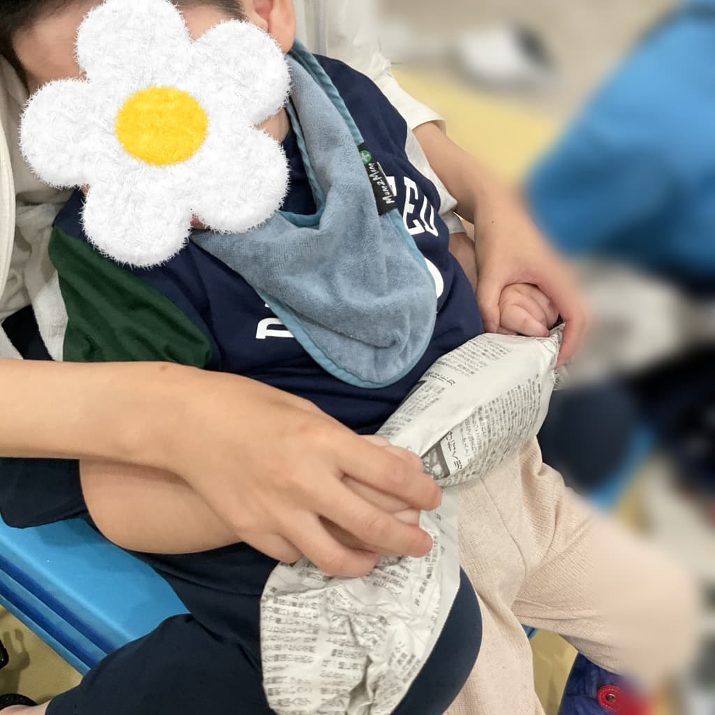 室内で、大人に身体を支えられた子どもが新聞紙を両手で持ち、膝の上に広げている様子