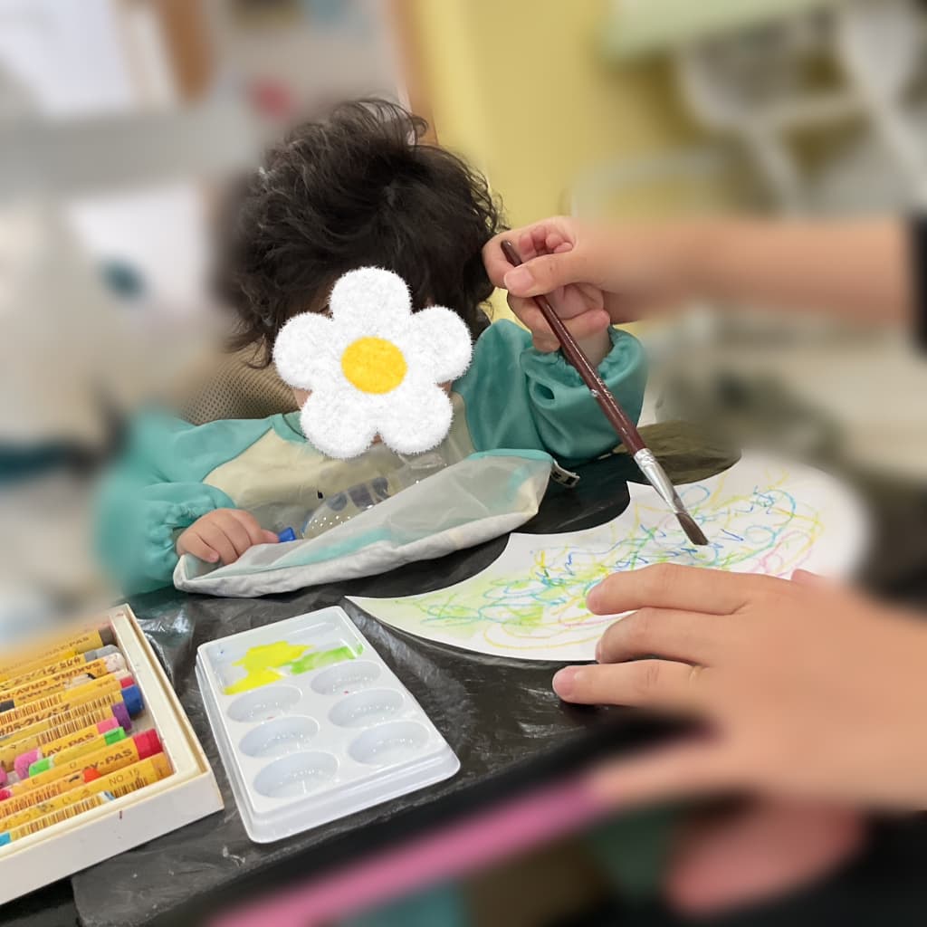 エプロンを着た子どもが、支援者に筆を支えてもらいながら、クレヨンや絵の具を使って円形の紙に自由に模様を描いている様子