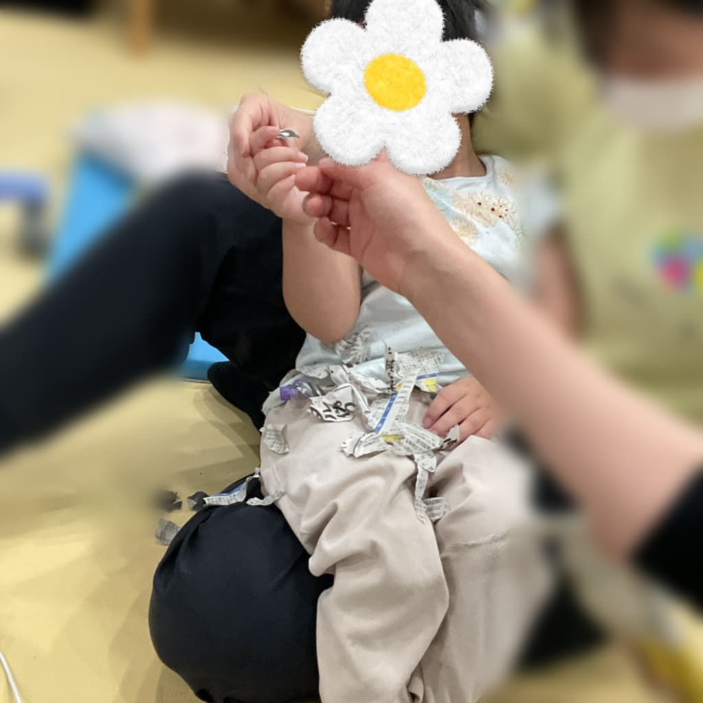 室内で、大人と向かい合った子どもが新聞紙のかけらを握っている。ひざの上や床に紙が広がっている様子