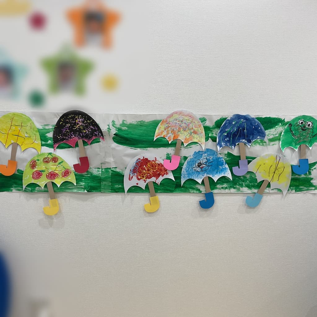 白い壁に、子どもたちが色を塗った傘の形の作品が横一列に飾られている様子。赤や青、黄色など色や模様が異なる傘が並び、梅雨をイメージした装飾になっている