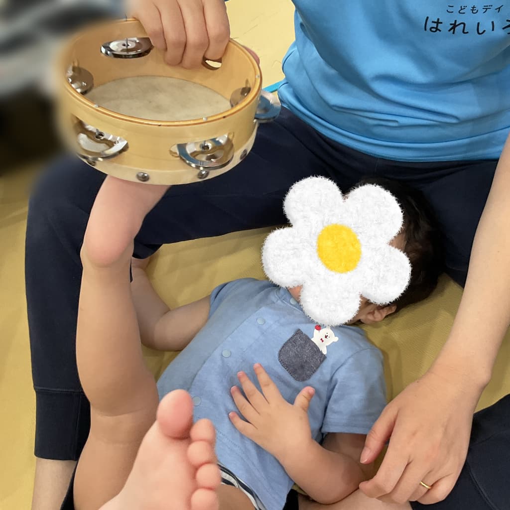 室内の床に敷かれたマットの上で、子どもが横になり、足を延ばして、支援者が持っているタンバリンに振れている様子