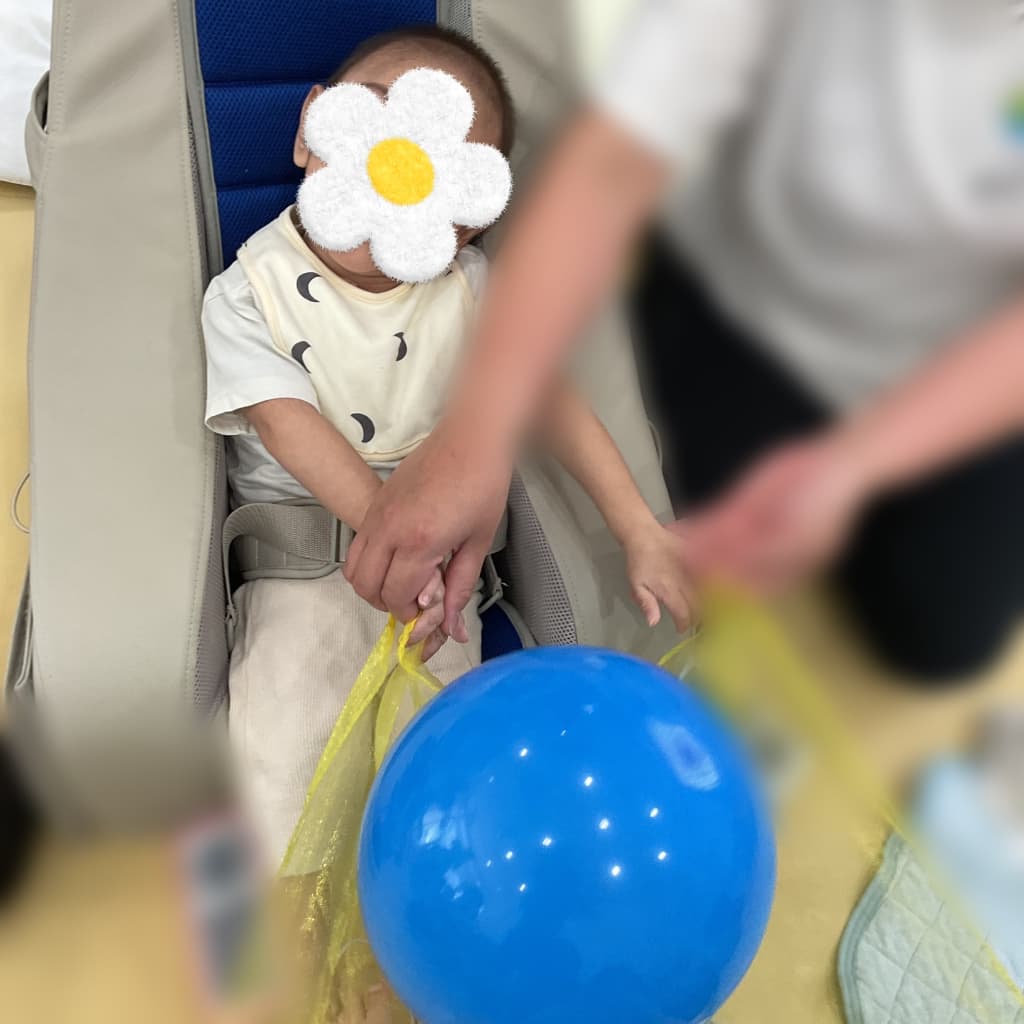 椅子に座った子どもが、支援者と一緒に黄色い薄布を持ち、その上で青い風船を動かしている様子