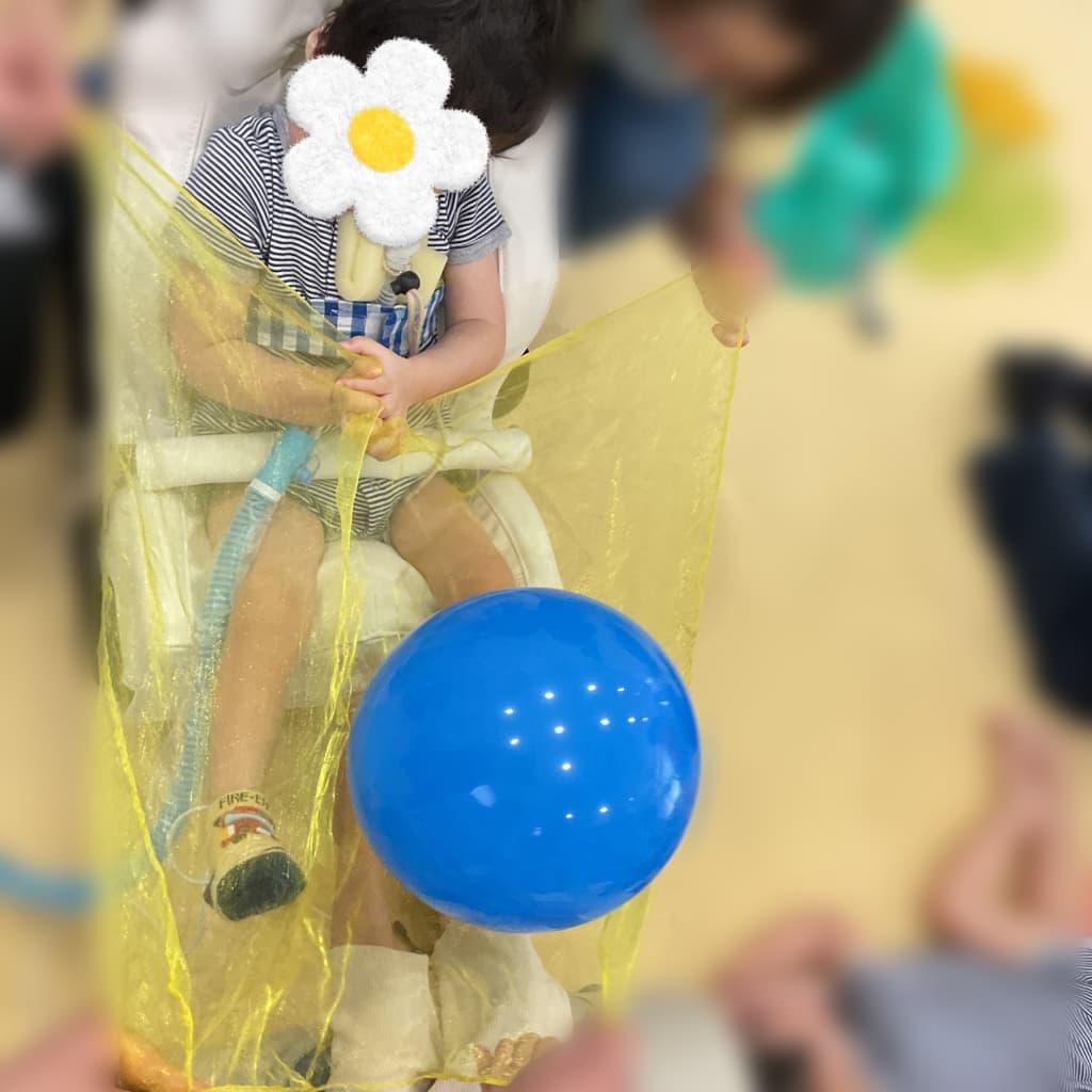 椅子に座った子どもの前に黄色い薄布を広げ、その上に青い風船を乗せて活動している様子