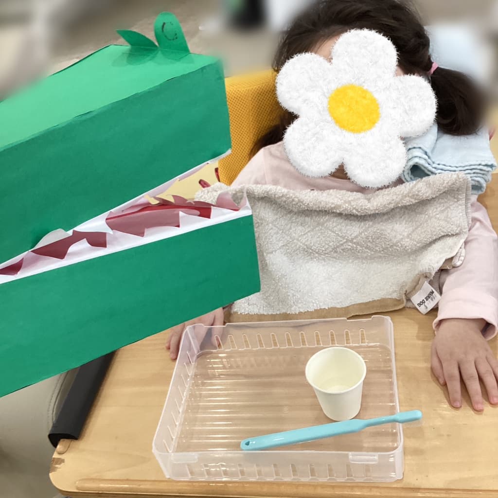 机の上に置かれた大きなワニの口の工作に向かい、子どもが椅子に座って活動に参加している様子。机の上にはトレーやコップ、おもちゃの歯ブラシが置かれている