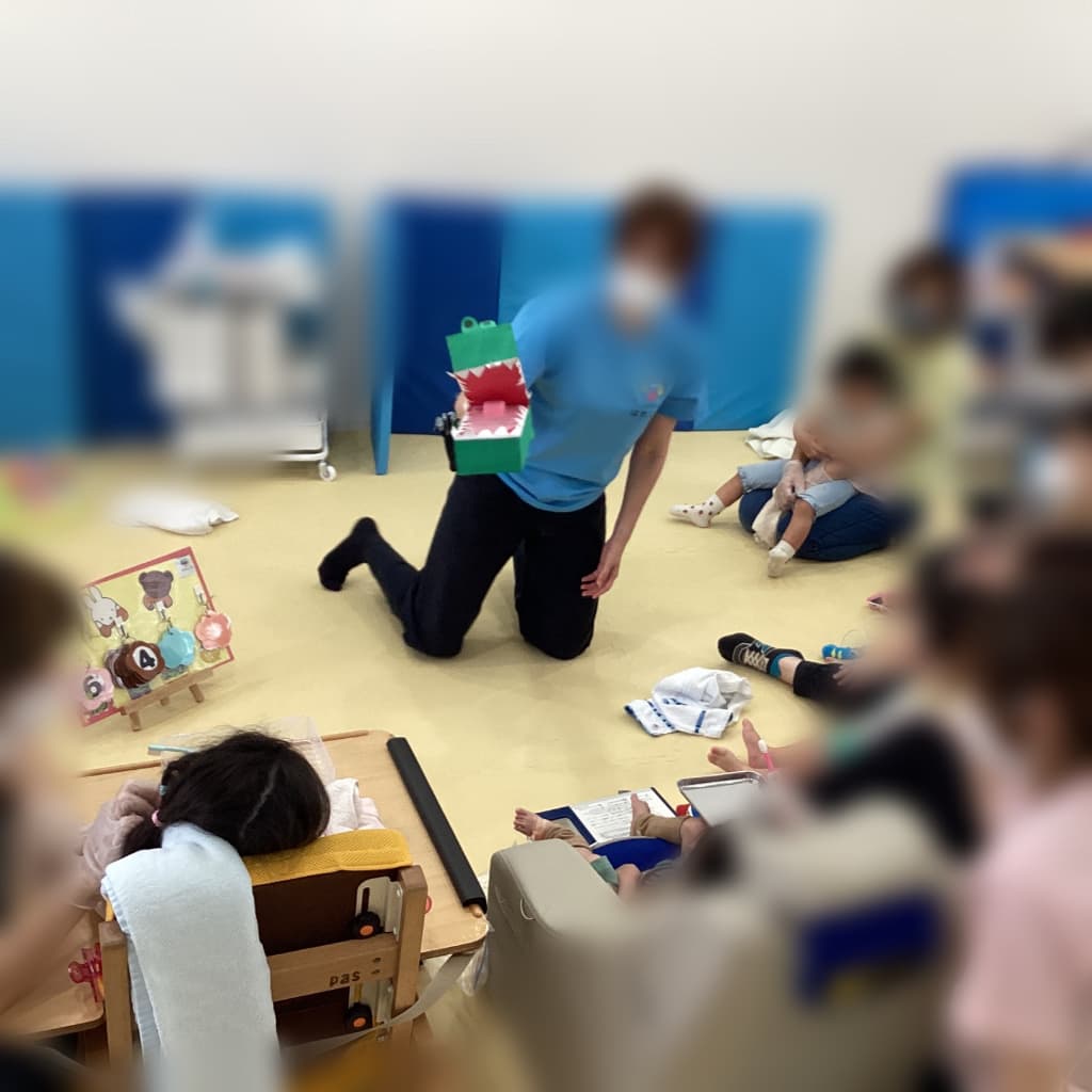 室内で、支援者がワニの工作を持ちながら中央に座り、周囲を囲むように子どもと大人が活動に参加している様子