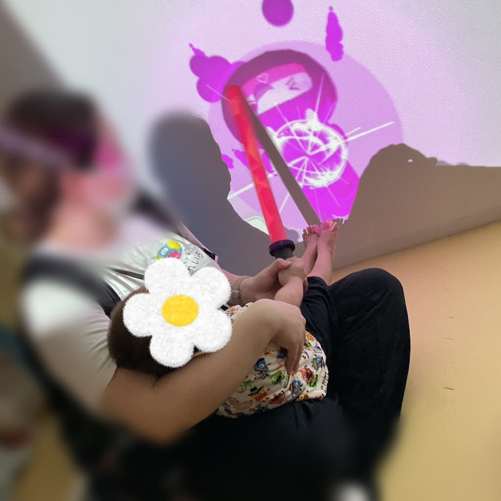 薄暗い室内で、大人に抱っこされた子どもが、壁いっぱいに映し出されたピンク色の映像を見ながら、赤いスティックを手に持っている様子