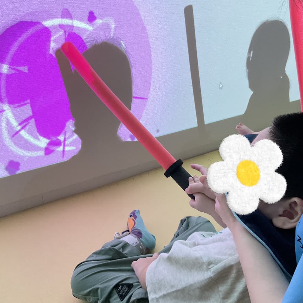 薄暗い室内で、座った子どもが赤いスティックを壁に向けて伸ばし、壁に映し出されたピンク色の映像と自分の影が重なっている様子