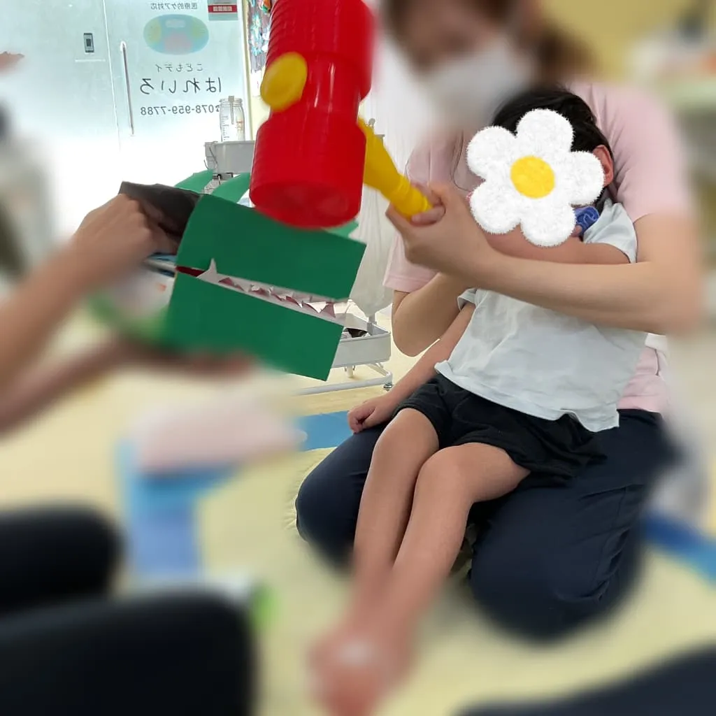 大人に抱っこされた子どもが、ハンマーのおもちゃで紙で作った緑のワニの模型をたたいている