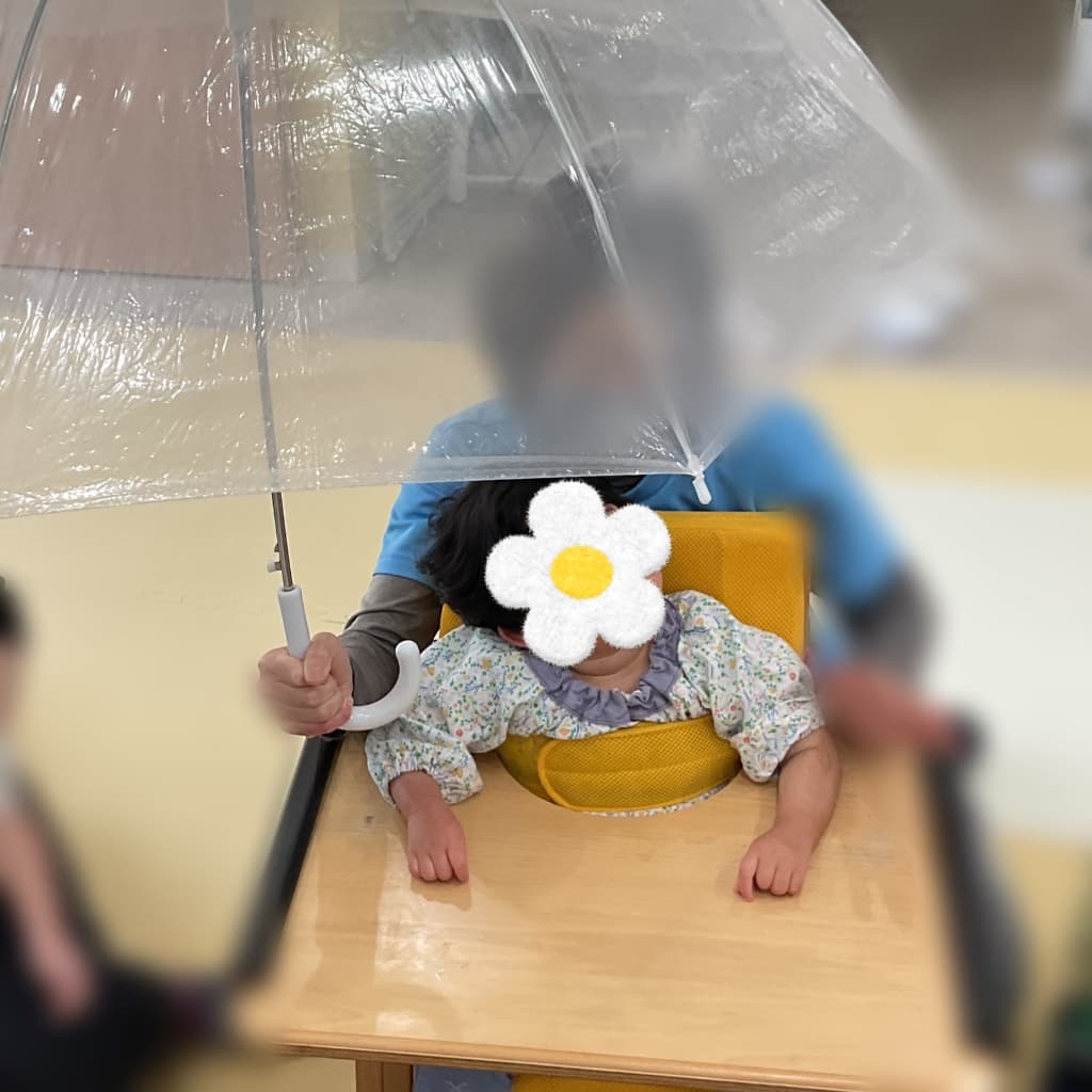 透明なビニール傘を支援者が子どもの頭上に広げ、椅子に座った子どもが傘を見上げている様子