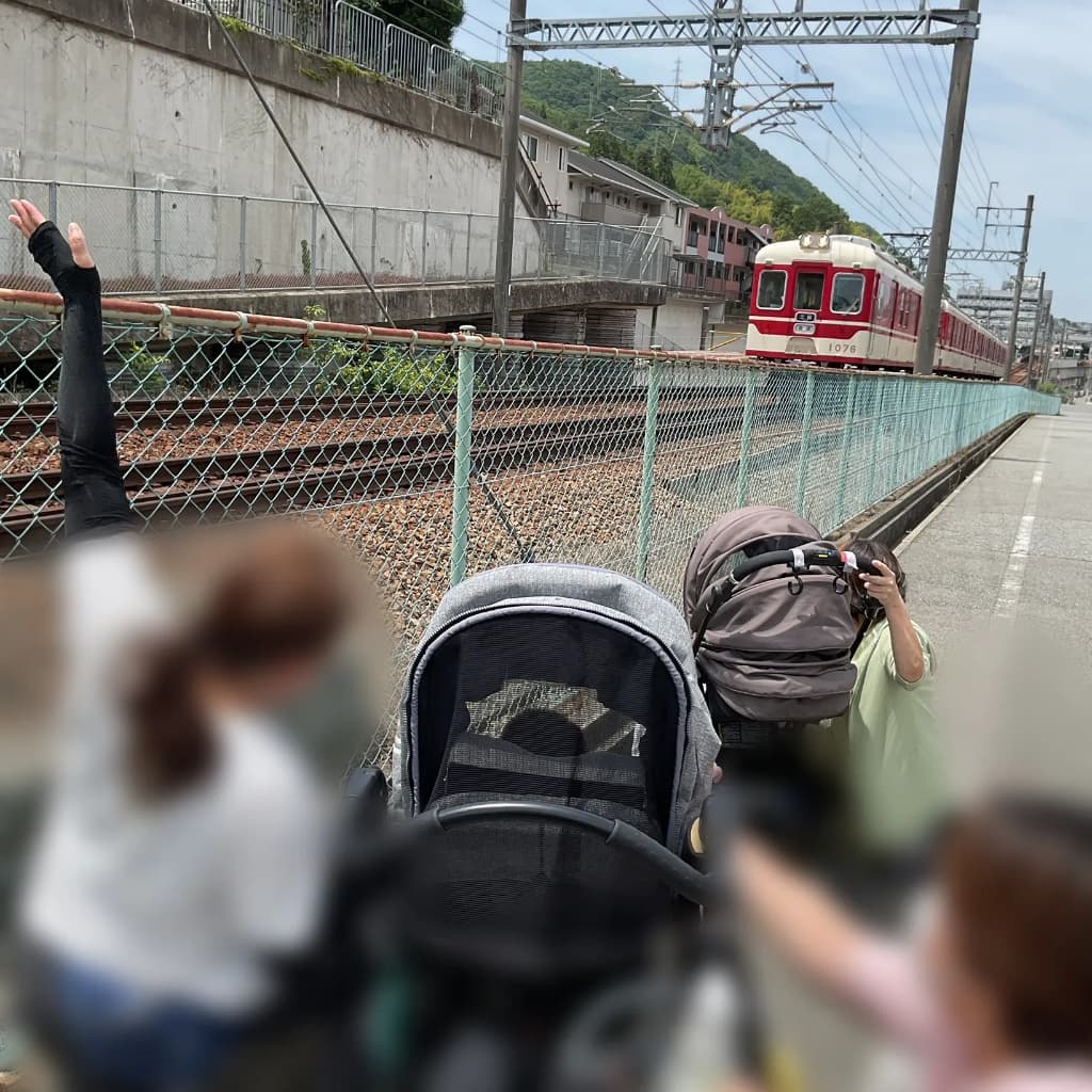道路わきを通過する列車に向けて、大人3人とベビーカーに乗った子どもが手を振っている