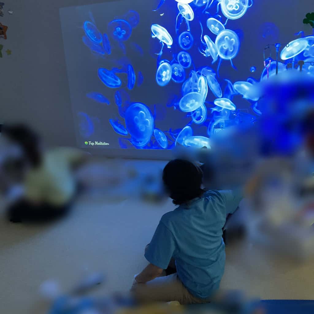 暗い部屋の壁に映し出されたクラゲの映像を座って眺めている子ども