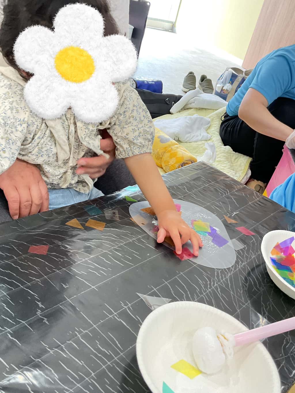 支援者に支えてもらいながら座り、制作をしている子ども。透明な丸いビニール台紙にカラフルなセロハンを張り付けている
