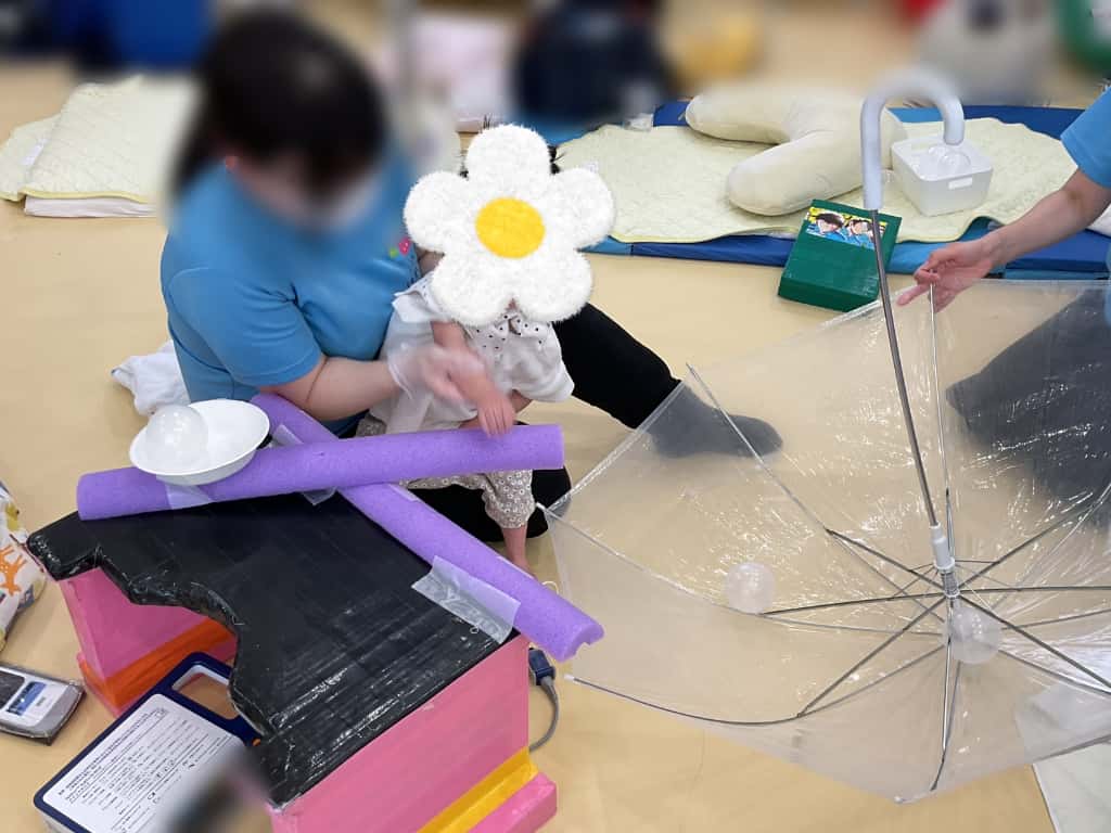 スポンジ製の棒を十字に組み合わせた先端に皿の付いた手作りの道具を使い、風船ボールを透明な傘の中へ入れようとしている様子