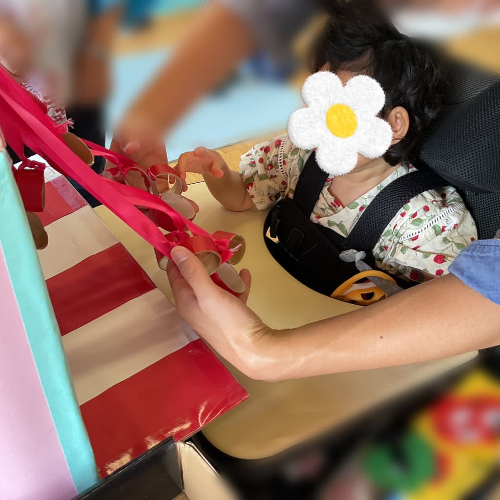 箱から垂れ下がる赤いひもに、車椅子に座った子どもが大人に支えてもらいながら手を伸ばしている