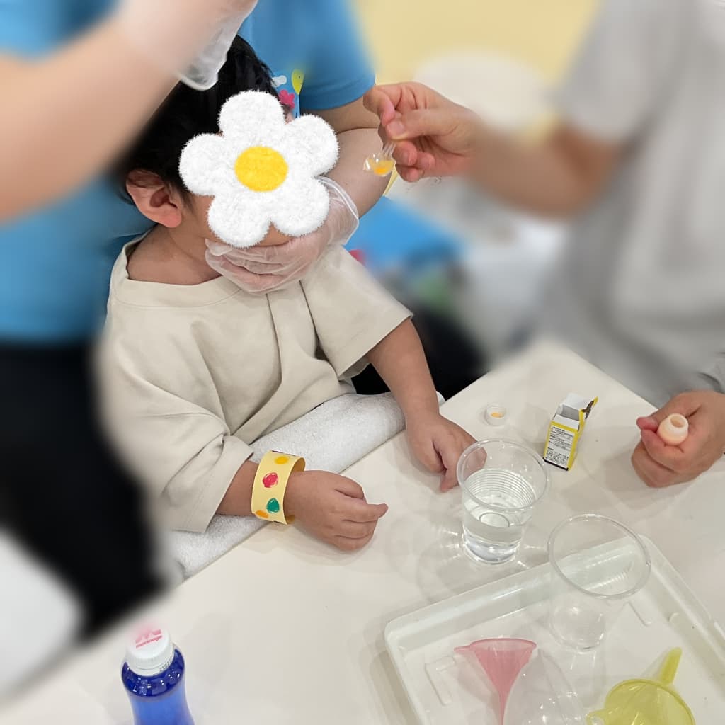 室内のテーブルでスタッフに抱っこしてもらいながら、食紅の色を選ぶ子ども