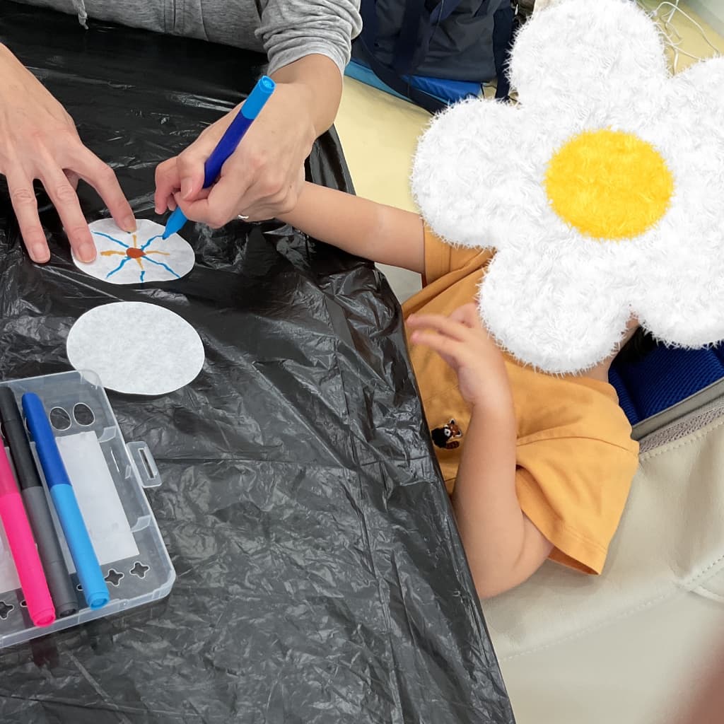 室内のテーブルでスタッフに手を支えてもらいながら、ペンで紙に模様を描く子ども