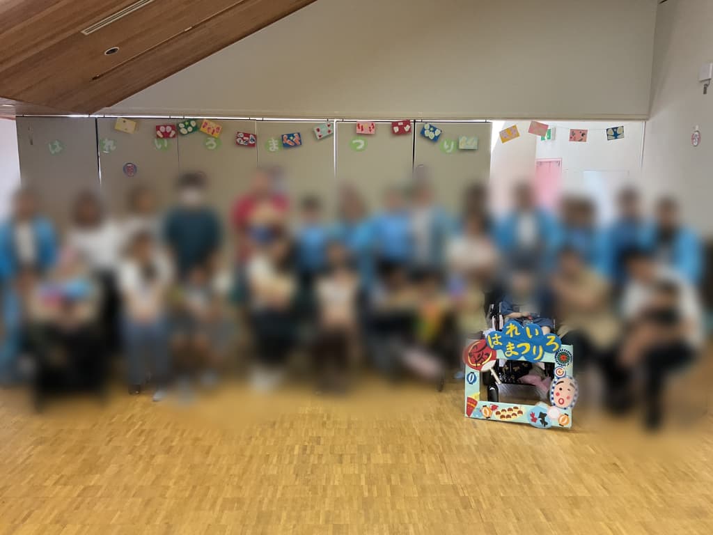 こどもデイはれいろ夏祭りで、参加者とスタッフが会場内に集まって撮影した集合写真。前方には「はれいろ夏まつり」と飾られたフォトスポットがある。
