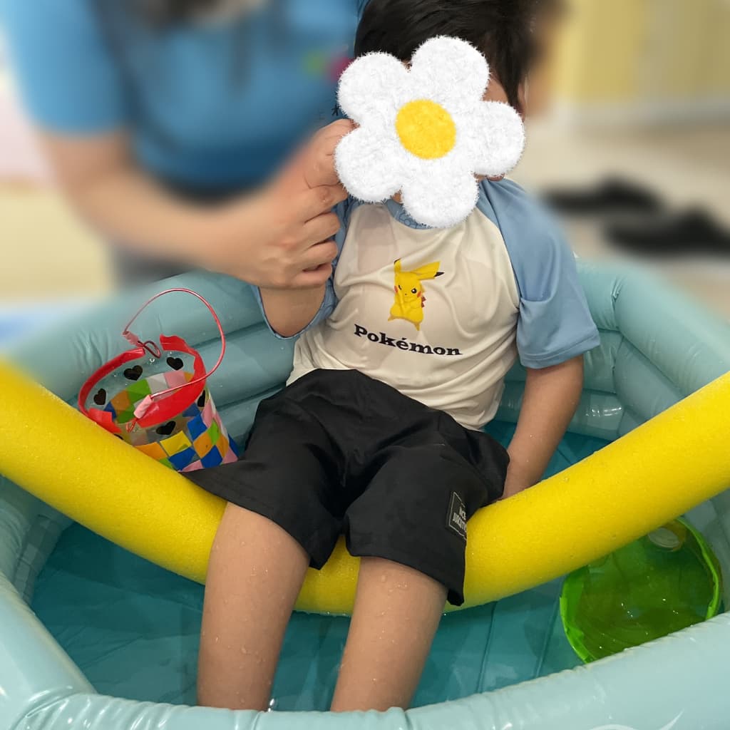 室内プールで大人に支えられながら、黄色い棒状のスポンジの上に座って、水に足をつけている子ども