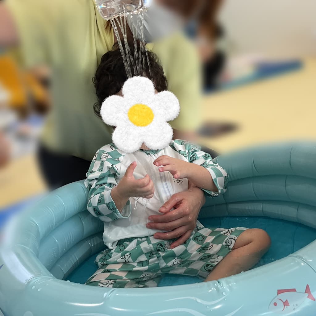 室内のビニールプールに座って遊んでいる子ども。上から大人がシャワーのように水を流している