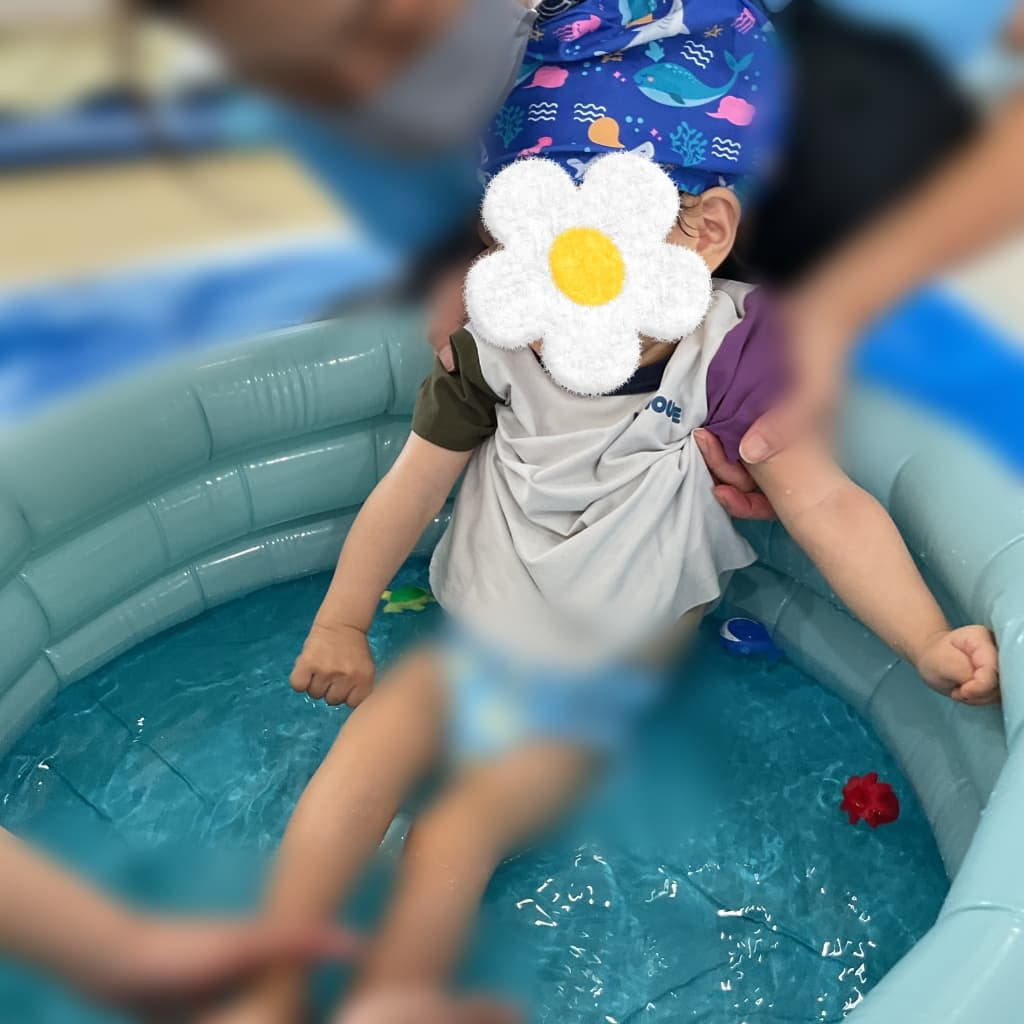 室内のビニールプールで大人に支えられながら水遊びをする子ども