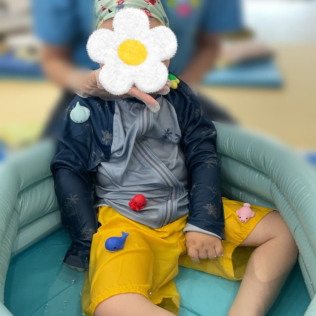 室内のビニールプールで大人に支えられながら水遊びをする子ども。タコやクジラのおもちゃに囲まれている