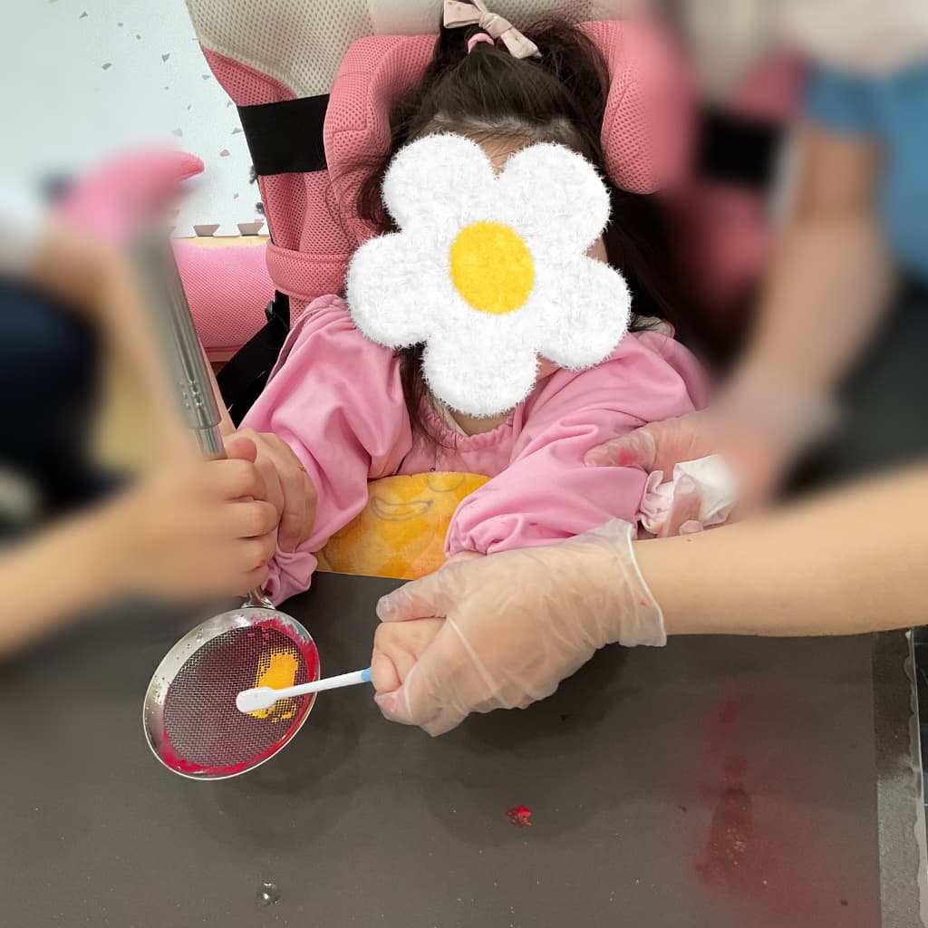 室内でスタッフに手を支えてもらいながら、ざるに絵の具のついた歯ブラシをこすりつけて制作をする子ども