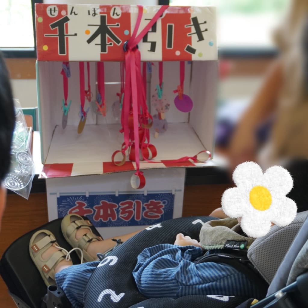 赤いひもと景品がたくさん吊り下げられた箱の前で、車椅子に座って遊ぶ子ども