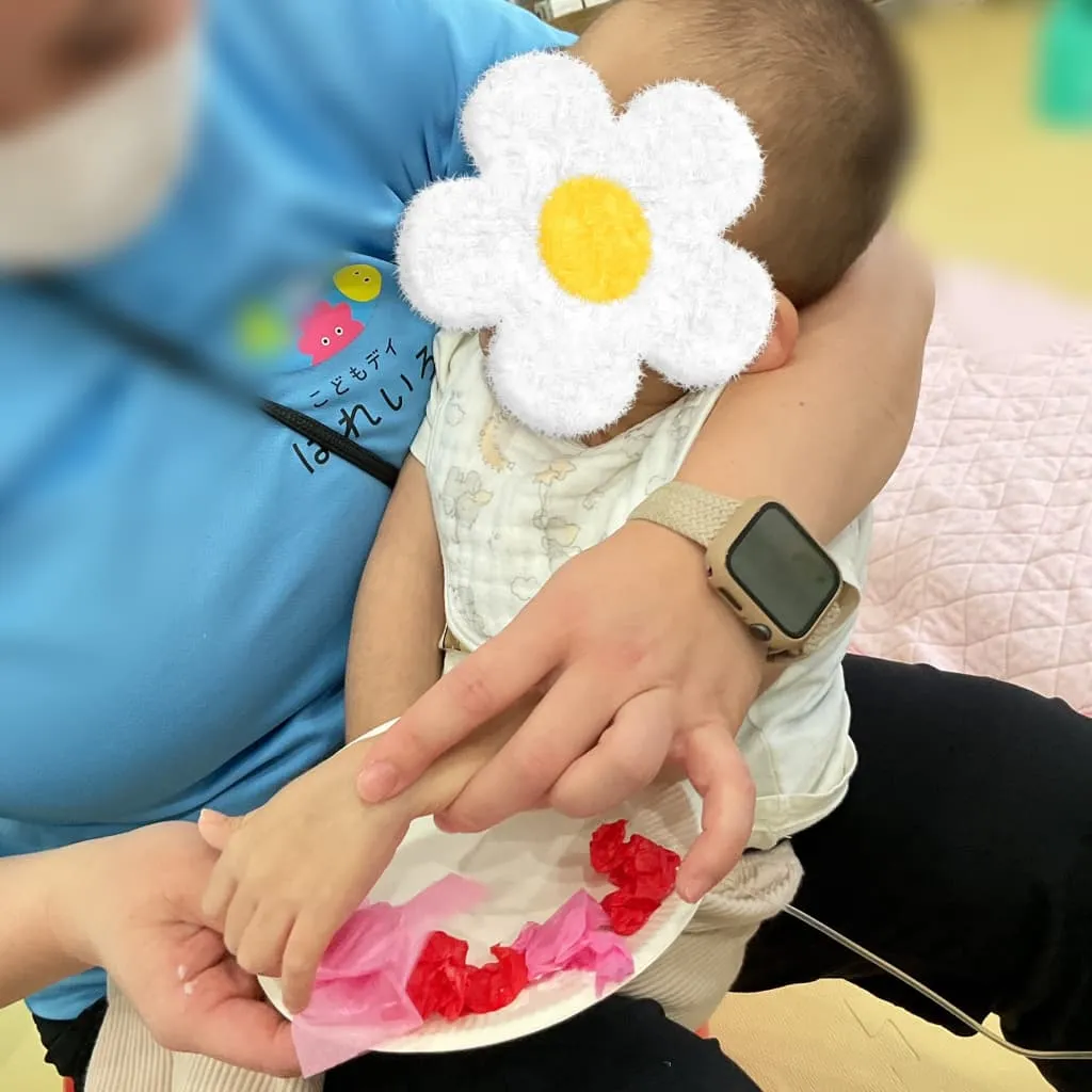 はれいろの支援員に抱っこされた乳児が、手を伸ばして赤やピンクの花紙に触れながら工作活動に参加している様子