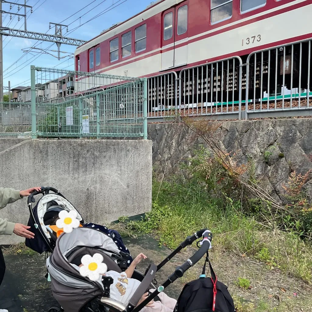 ベビーカーに乗った子どもたちが線路沿いで赤い電車を見上げなている様子