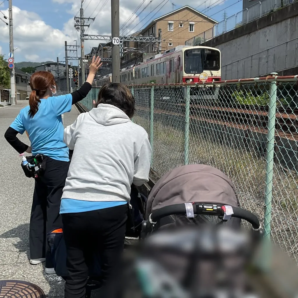 支援員2人とベビーカーに乗った子どもたちが線路沿いで通過する電車に向かって手を振っている様子
