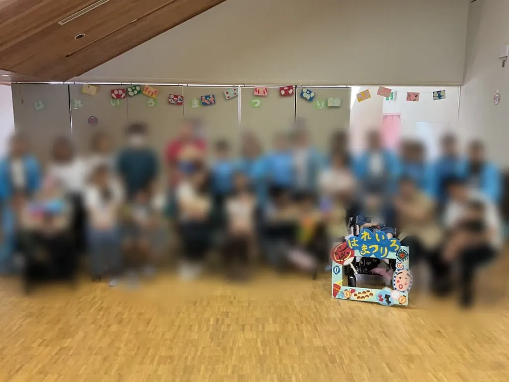 こどもデイはれいろ夏祭りで、参加者とスタッフが会場内に集まって撮影した集合写真。前方には「はれいろ夏まつり」と飾られたフォトスポットがある。