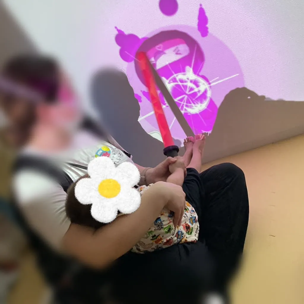 薄暗い室内で、大人に抱っこされた子どもが、壁いっぱいに映し出されたピンク色の映像を見ながら、赤いスティックを手に持っている様子